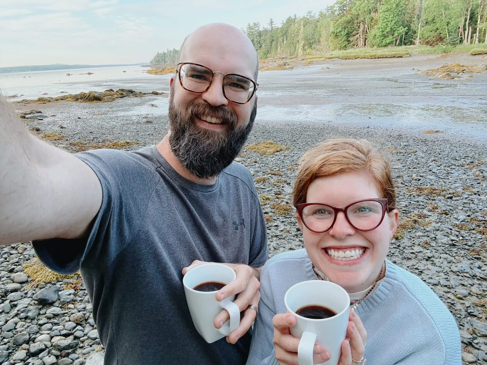 Jake and a friend smiling with mugs of coffee on a rocky shoreline