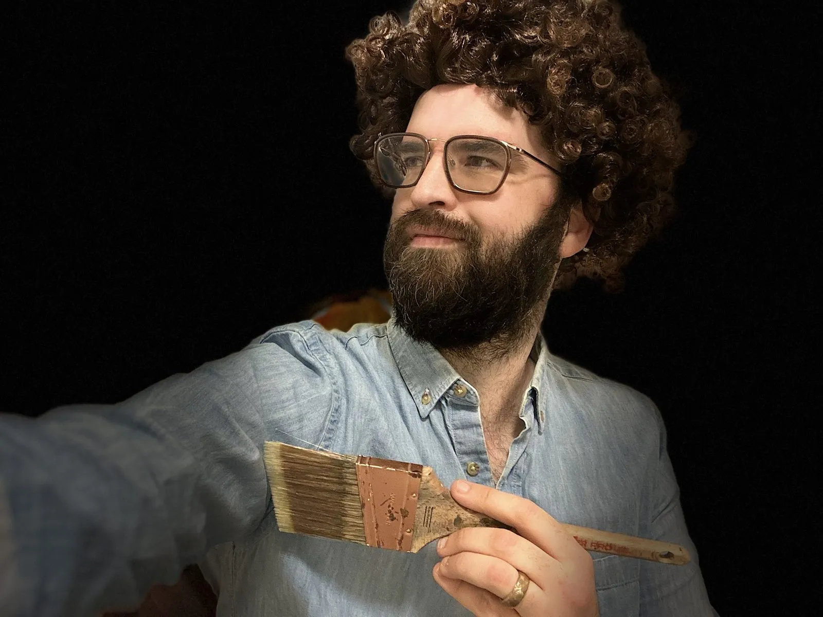 Jake in a curly wig holding up a paintbrush, Bob Ross style