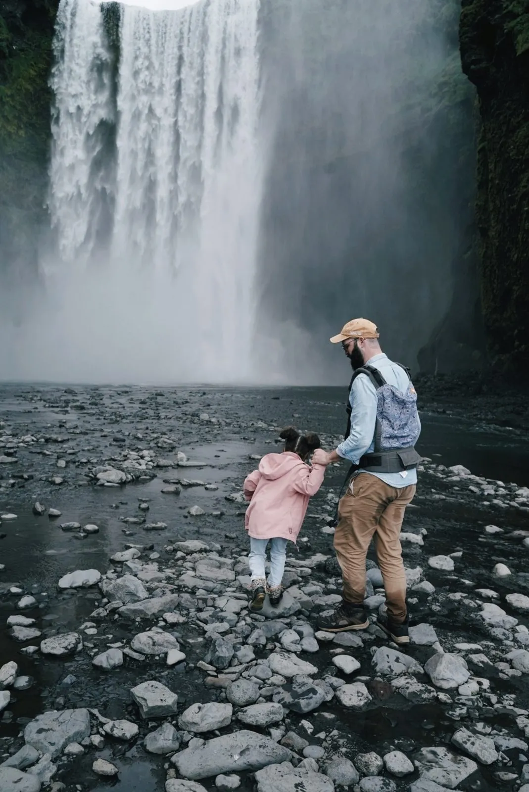 Jake holding a small child's hand, walking over rocks toward a huge waterfall